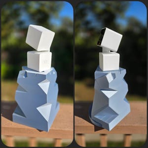 May include: A modern sculpture featuring a light blue geometric base with a white, cube-shaped structure on top. The sculpture has a minimalist design, with clean lines and a contemporary aesthetic. The background is blurred, suggesting an outdoor setting.
