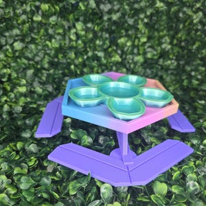 May include: A colorful, tiered serving tray with six teal-colored bowls. The tray has a gradient of colors, transitioning from blue to pink. The legs are purple, and the background is a dense green foliage.