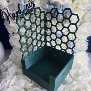 May include: A teal-colored, honeycomb-patterned display stand with the text "Hayden's Minis". The stand has a rectangular base and a back with a geometric design. The background features white and light blue flowers.