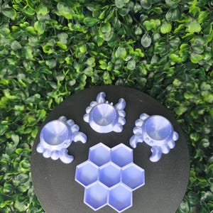 May include: Three light purple crab-shaped stands with circular indentations and one light purple hexagonal honeycomb-shaped tray with seven individual compartments, all arranged on a circular black surface.