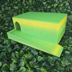 May include: A rectangular green and yellow gradient plastic enclosure featuring a small arched entrance, a textured ramp, and embossed beetle illustrations on the side. The structure has a flat top and a ribbed base, designed with a smooth finish.