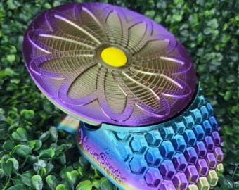 May include: A 3D printed object with a circular top featuring a purple and gold flower design with a yellow center. The base has a blue, purple, and gold iridescent honeycomb texture. The text "Hovr 3D Minis" is embossed on the side of the base.