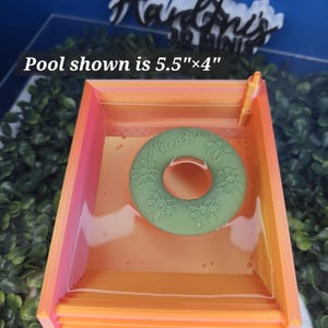 May include: A rectangular orange and pink pool measuring 5.5 inches by 4 inches, containing a green circular float with a floral pattern and the text Hardin's. The pool features a small orange post in one corner and a clear liquid interior.