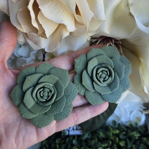 May include: Two sage green succulent-shaped ornaments featuring a layered, petal-like structure with visible 3D-printed horizontal striations on the surface of each leaf. The objects are circular in form, mimicking the appearance of a blooming plant.