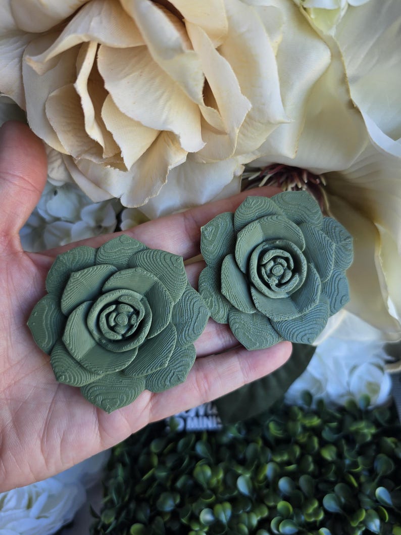 May include: Two sage green succulent-shaped ornaments featuring a layered, petal-like structure with visible 3D-printed horizontal striations on the surface of each leaf. The objects are circular in form, mimicking the appearance of a blooming plant.