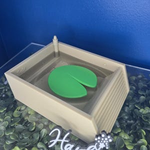 May include: A beige rectangular pool structure with a single green lily pad floating in the center. The pool features a small pillar in one corner, a set of steps on one side, and the word "Sale" embossed on the inner wall. A white logo reads "Hardinis 3D MINIS".