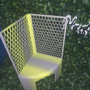 May include: A small, white, corner chair with a honeycomb pattern on the back and side. The chair is made of a light-colored material and has a simple, modern design. The chair is set against a backdrop of green foliage.