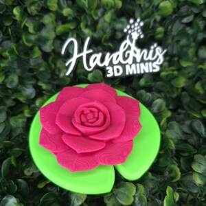 May include: A bright pink 3D printed rose flower resting on a smooth, circular, lime green lily pad. Above the flower, a black sign with white text reads Hardini's 3D MINIS, featuring a small star-shaped graphic above the letter i.