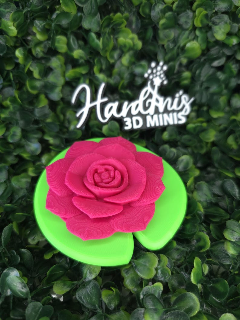 May include: A bright pink 3D printed rose flower resting on a smooth, circular, lime green lily pad. Above the flower, a black sign with white text reads Hardini's 3D MINIS, featuring a small star-shaped graphic above the letter i.