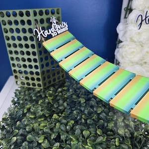 May include: A green rectangular tower with a grid pattern attached to a suspended bridge made of six rectangular planks featuring a yellow, light green, and teal gradient. A white sign with the text Hardni's 3D MINIS is attached to the bridge.