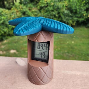 May include: A brown palm tree-shaped weather station with a digital display showing the temperature as 84.4 degrees Fahrenheit and the humidity as 49%.