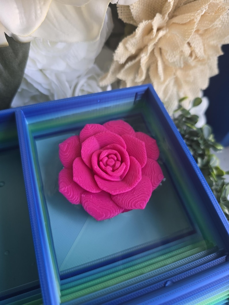 May include: A vibrant hot pink 3D printed rose with visible layered texture, centered inside a square blue and green gradient frame. The flower features tightly packed petals arranged in a circular pattern, resting on a flat, light blue base.