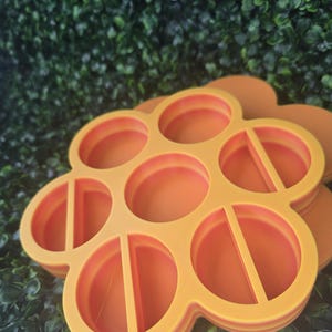 May include: A bright orange, flower-shaped plastic tray featuring seven circular compartments. The central compartment is open, while the six surrounding outer compartments each contain a single vertical divider bar across the center.