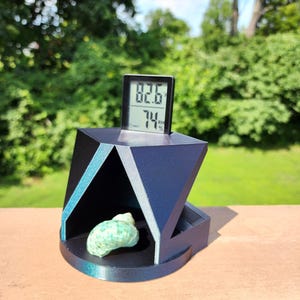 May include: A geometric, dark blue display stand with a digital thermometer showing 82.6 degrees Fahrenheit and 74% humidity. A small, green seashell rests inside the stand. The stand has teal accents.