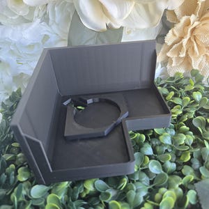 May include: A matte black L-shaped corner shelf featuring a central circular cutout designed for mounting or holding a specific object. The shelf has a flat base and two perpendicular back walls, constructed with a smooth, uniform finish.