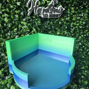 May include: A 3D printed display stand featuring a gradient color scheme transitioning from light green at the top to dark blue at the base. The stand has a square back wall with the embossed text Hardini's and a curved front opening.
