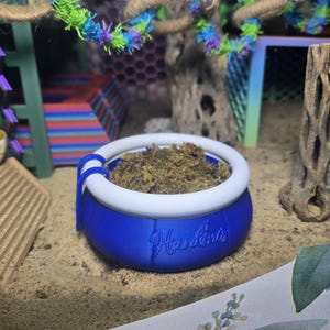 May include: A blue and white pet habitat accessory with the word "Hardin's" on the side. The bowl is filled with brown moss. The background includes a decorative tree branch with colorful accents.