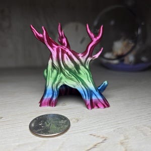 May include: A 3D printed tree-shaped object with a gradient finish transitioning from pink at the top to green in the middle and blue at the base. The object is positioned next to a US quarter coin for scale, showing its small size and textured surface.