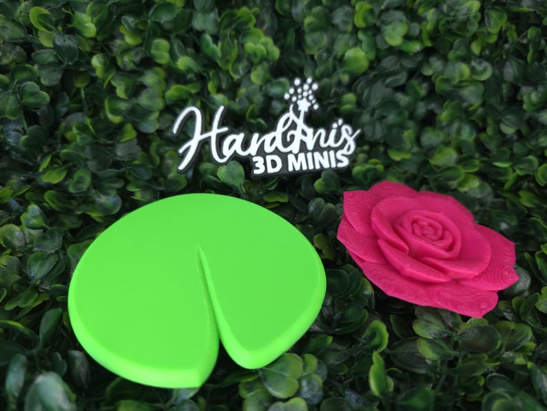 May include: A bright green lily pad and a vibrant pink rose flower figurine. Above the items, a black sign with white text reads HardAni's 3D MINIS with a small star graphic. The objects are smooth and appear to be made of a solid material.