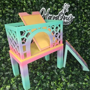 May include: A colorful, multi-tiered pet habitat with a ramp. The structure features a cage-like design with geometric cutouts, a yellow interior, and a rainbow gradient of colors including pink, yellow, and teal. The habitat sits on a green leafy background.