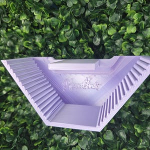 May include: A lavender-colored, triangular ashtray with a stepped design on the sides. The ashtray has a small platform and the word "Handmade" is visible. The ashtray is set against a backdrop of green foliage.
