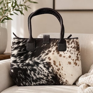 May include: A tote bag featuring a black and white cowhide pattern with brown spots. The bag has two dark brown leather handles, a top zipper closure, and a central leather tab with a snap button fastener on the front exterior.