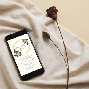 May include: A black smartphone displays a digital invitation for a Christmas party. The invitation features a gold circular frame with green leaves and the German text "Weihnachtsfeier". The phone rests on a beige fabric with dried plant stems.