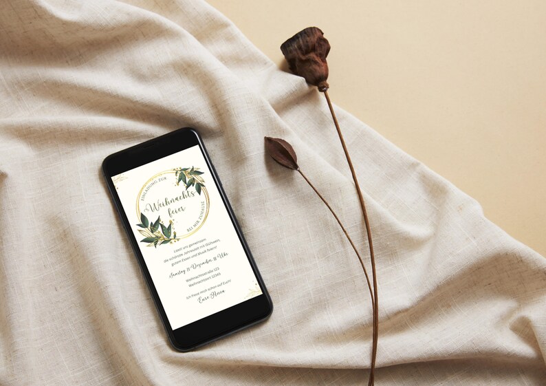 May include: A black smartphone displays a digital invitation for a Christmas party. The invitation features a gold circular frame with green leaves and the German text "Weihnachtsfeier". The phone rests on a beige fabric with dried plant stems.