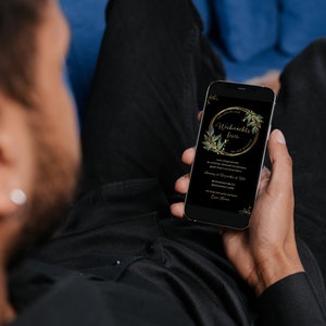 May include: A smartphone displays a black screen with gold text and a gold and green floral design. The text reads "Weihnachts feier" in a gold oval. The phone is held by a person wearing a black shirt, sitting on a blue couch.