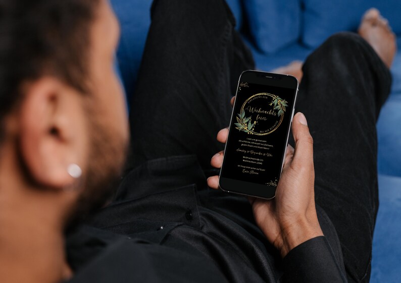 May include: A smartphone displays a black screen with gold text and a gold and green floral design. The text reads "Weihnachts feier" in a gold oval. The phone is held by a person wearing a black shirt, sitting on a blue couch.