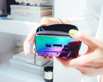 May include: A small, iridescent cassette tape-shaped wallet with a black zipper and a silver zipper pull. The wallet is held open, revealing a black interior. The colors shift from green to purple. The background is a white wall and a white shelf.