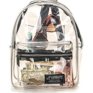 May include: Clear, transparent backpack with a black zipper and straps. The backpack contains sunglasses, cash, keys, and a lip balm. A small black label reads "FYDELITY BAG COMPANY, BROOKLYN, CALIFORNIA, ESTABLISHED 2009."