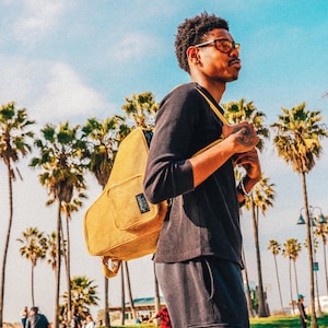 May include: A person wearing a black long-sleeved shirt and dark grey sweatpants with a yellow corduroy backpack. The person is standing in front of a palm tree and a blue sky with white clouds. The text "CARRY EXTRA VIBES" is at the top of the image.