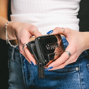 May include: A black cassette tape-shaped wallet with a zipper closure and a silver metal accent. The wallet is being held by a person with blue nail polish.