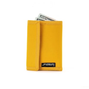 May include: Mustard yellow wallet with a flap closure, holding two one-hundred dollar bills. The wallet has a rectangular shape and a small black label with white text that reads "FIDELITY".