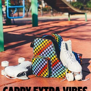 May include: A rainbow checkered roller skate backpack with a white roller skate inside. The backpack has a small front pocket. The text "Carry Extra Vibes" is printed below the image.
