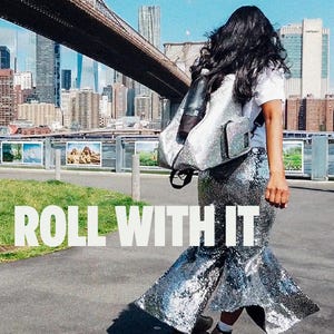 May include: A person wearing a sequined silver skirt and roller skates, with a silver backpack, stands on a paved path. The text "ROLL WITH IT" is displayed in white block letters. The background features a bridge and city skyline.
