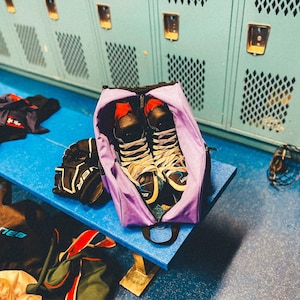 May include: Purple sports bag with black straps and a zipper. The bag is open and contains two pairs of black ice skates with white and blue accents. The bag is sitting on a blue bench in front of a locker room.