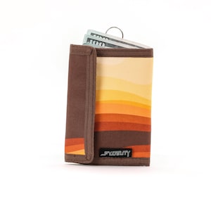 May include: A tri-fold wallet with a retro sunset design in shades of orange, yellow, and brown. The wallet is open, revealing a few folded bills and a metal key ring. The brand name "FIDELITY" is printed on the front.