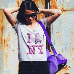 May include: A woman wearing a white t-shirt with a pink "I ♥ NY" graphic featuring a roller skate. She is also wearing black jeans and sunglasses. She is carrying a purple corduroy tote bag with a black strap.