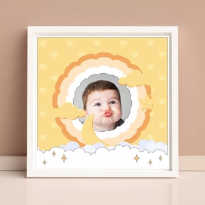 Puede incluir: Una impresión enmarcada amarilla y blanca con la cara de un bebé en un círculo rodeado de nubes y estrellas. El bebé está haciendo una mueca divertida.
