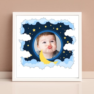 Puede incluir: Una impresión enmarcada con un retrato de bebé dentro de un diseño de cielo nocturno. La imagen incluye un círculo azul con la cara de un bebé, rodeado por un fondo azul oscuro con estrellas amarillas, nubes blancas y una luna creciente amarilla.
