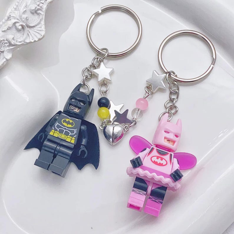 May include: A pair of keychains featuring Lego minifigures of Batman and Batgirl. The keychain is decorated with silver stars, a heart charm, and pink and clear beads.
