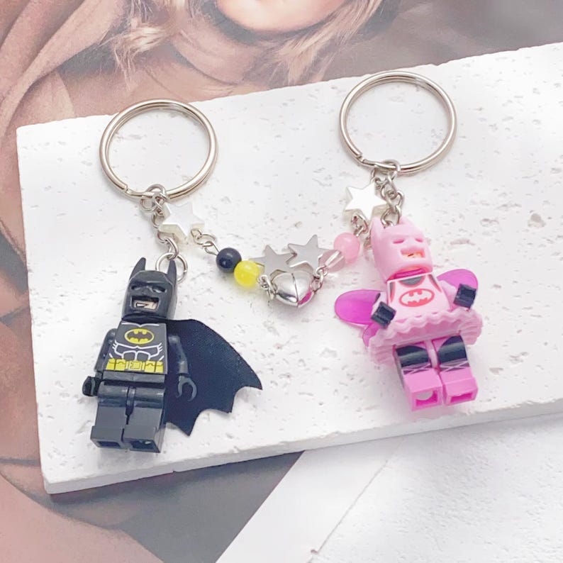May include: A pair of keychains featuring a black and yellow Batman Lego minifigure and a pink and black Batman Lego minifigure with a pink tutu and wings. The keychains are connected by a silver chain with black and yellow beads and silver stars.