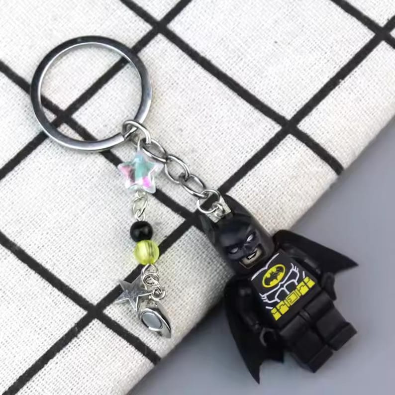May include: A black Batman Lego minifigure keychain with a yellow bat symbol on the chest. The keychain has a silver ring, a silver chain, and a silver star charm with iridescent glitter. The keychain also has a black and yellow bead charm.