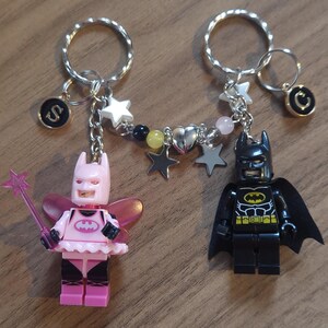 May include: Two Lego minifigure keychains, one dressed as a pink fairy Batman and the other as a traditional black Batman. The keychains are connected by a silver chain with charms, including stars, hearts, and beads.