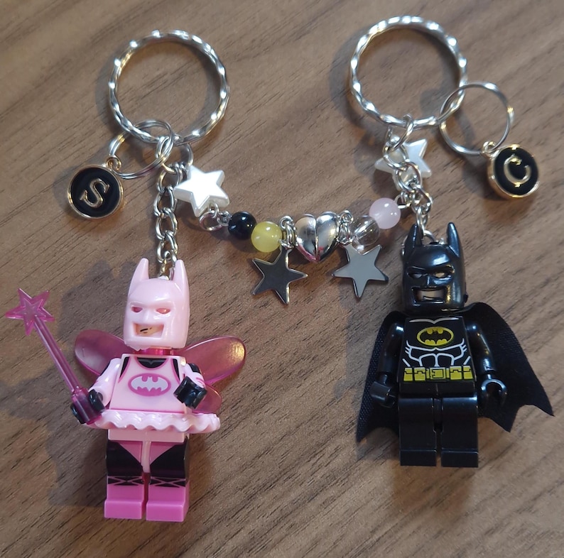 May include: Two Lego minifigure keychains, one dressed as a pink fairy Batman and the other as a traditional black Batman. The keychains are connected by a silver chain with charms, including stars, hearts, and beads.