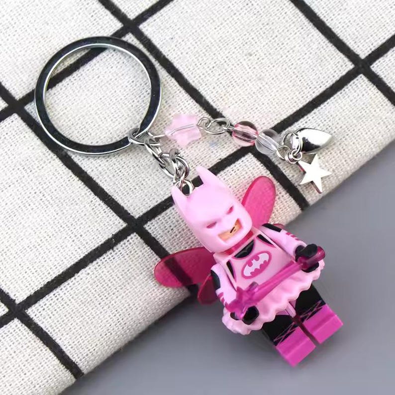 May include: Pink Batman fairy keychain with silver wings, a silver key ring, and a silver charm with a star. The keychain has pink and black details and a silver charm with a heart.