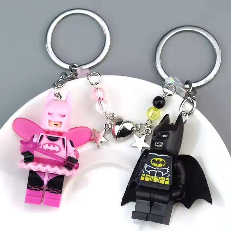 May include: A pink and black Lego minifigure keychain with a silver chain and charms. The pink minifigure is a female character with wings and a wand. The black minifigure is Batman.