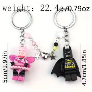 May include: A pink and black Lego minifigure keychain with a silver chain and star charms. The pink minifigure is wearing a pink dress and wings. The black minifigure is Batman. The keychain is 5 inches long and weighs 22.4 grams.